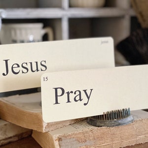 May include: Two cream-coloured cards with the words "Jesus" and "Pray" printed in black. The cards are propped up on a stack of old books. The word "Jesus" is above the number 4, and "Pray" is above the number 15.