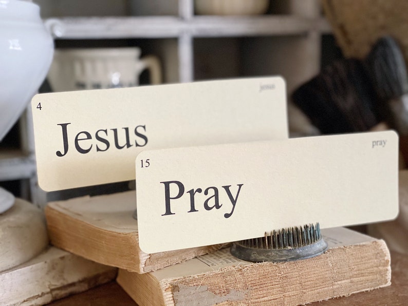May include: Two cream-coloured cards with the words "Jesus" and "Pray" printed in black. The cards are propped up on a stack of old books. The word "Jesus" is above the number 4, and "Pray" is above the number 15.