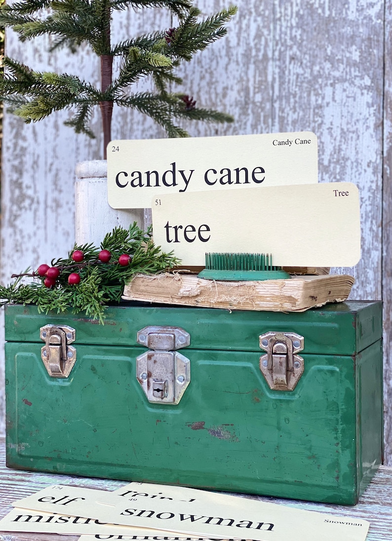Peut inclure: Une bo&icirc;te &agrave; outils en m&eacute;tal vert vintage avec des fermoirs argent&eacute;s contient des cartes de mots sur le th&egrave;me des f&ecirc;tes. Un petit sapin de No&euml;l et des brins de verdure avec des baies rouges sont pos&eacute;s sur la bo&icirc;te &agrave; outils, cr&eacute;ant une sc&egrave;ne de vacances.