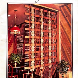 May include: A macrame wall hanging with a wood frame and tassels. The hanging is made with natural colored cord and is hung in a room with wood paneling.