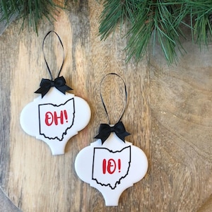 May include: Two white ceramic ornaments with a black outline of the state of Ohio and the letters "OH!" in red. Each ornament has a black ribbon and bow for hanging. The ornaments are on a wooden surface.