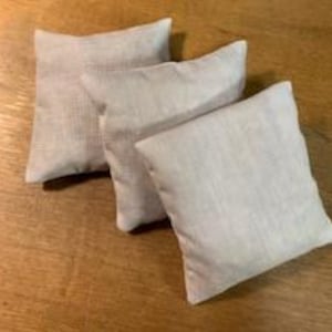 May include: Three small, square, light gray linen pillows arranged on a wooden surface. The pillows have a textured appearance and are slightly different sizes. The lighting creates subtle shadows, enhancing the fabric's texture.