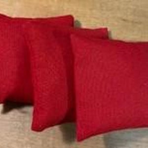 May include: Three red bean bags are arranged on a wooden surface. The bean bags are square-shaped and appear to be made of a textured fabric. The lighting highlights the texture of the fabric and the vibrant red color of the bags.