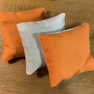 May include: Three small, square bean bags. Two are bright orange, and one is light gray. The bean bags are made of a textured fabric and are arranged on a wooden surface. Each bag is approximately 6 inches square.