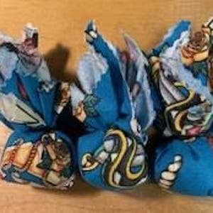 May include: Three small, round fabric bundles with a teal background and various colorful illustrations. The illustrations include a snake, a horse, and roses. The bundles are tied with a lighter fabric, creating a decorative, flower-like appearance.