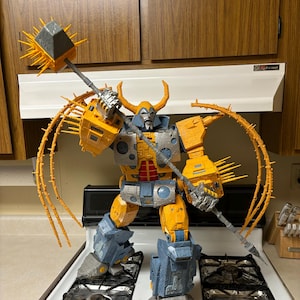 Puede incluir: Una figura de acción grande y articulada en tonos amarillos, grises y azules, sosteniendo una maza con púas. La figura tiene detalles intrincados, incluyendo cuernos y apéndices puntiagudos. La figura está sobre una estufa.