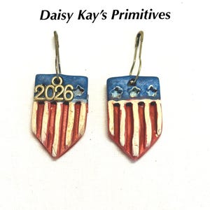 May include: Patriotic shield-shaped earrings with red and white stripes and a blue top with stars. The earrings have a metal hook and the year "2026" is displayed on one earring. The text "Daisy Kay's Primitives" is above the earrings.