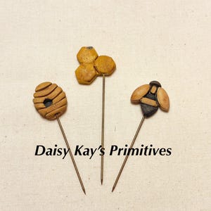May include: Three handcrafted pins with unique designs: a beehive, a honeycomb, and a bee. Each pin features a detailed, textured design in shades of brown and tan, mounted on a long, slender, gold-colored pin. The text "Daisy Kay's Primitives" is below the pins.