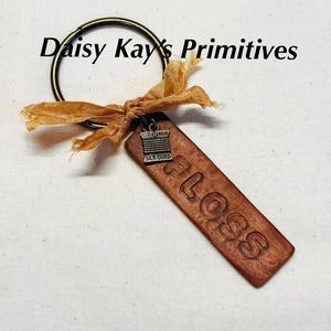 May include: A keychain featuring a rectangular wooden tag embossed with the word FLOSS, a small metal charm shaped like a spool of thread labeled SILK CURD, and a bronze-toned metal ring tied with a frayed mustard-yellow fabric ribbon.