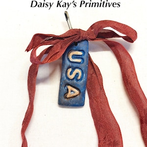 May include: A blue rectangular tag with the letters "USA" in gold, attached with a safety pin and tied with a red ribbon. The tag is handmade and has a rustic appearance. The text "Daisy Kay's Primitives" is at the top.