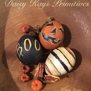 May include: A wooden bowl filled with Halloween-themed decorations. The decorations include three painted gourds: one black with "BOO" in gold, one orange carved like a jack-o'-lantern, and one with black, orange, and yellow stripes. Small orange pumpkins and dried leaves are also present. The text "Daisy Kays Primitives" and "Kristy Green" are visible.