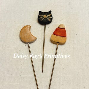 May include: Three decorative pins with wooden toppers. One is a crescent moon, another is a black cat with a gold bow, and the third is a candy corn design. The pins are on a light-colored background. The text "Daisy Kay's Primitives" is also visible.