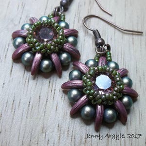 May include: Handmade earrings with a floral design. Each earring features a central faceted gem surrounded by a ring of green beads. The petals are a muted purple, and the outer beads are a silvery-blue color. The earrings have a hook closure.
