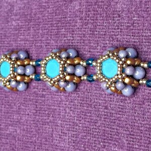 K&ouml;nnte beinhalten: Ein Armband mit Perlen und einem goldenen Verschluss. Das Armband hat t&uuml;rkisfarbene, goldene und lavendelfarbene Perlen, die in einem Blumenmuster angeordnet sind. Das Armband liegt auf einem lila Hintergrund.