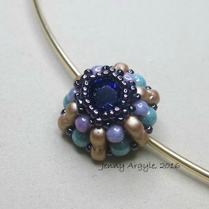 K&ouml;nnte beinhalten: Eine Halskette mit Anh&auml;nger aus Perlen, mit einem dunkelblauen Mittelstein, umgeben von t&uuml;rkisfarbenen, lavendelfarbenen und beigefarbenen Perlen. Der Anh&auml;nger ist an einem goldfarbenen Draht befestigt. Der Text "Jenny Argyle, 2016" ist sichtbar.