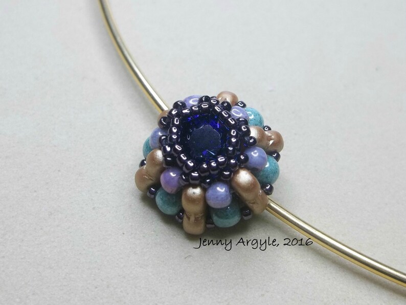K&ouml;nnte beinhalten: Eine Halskette mit Anh&auml;nger aus Perlen, mit einem dunkelblauen Mittelstein, umgeben von t&uuml;rkisfarbenen, lavendelfarbenen und beigefarbenen Perlen. Der Anh&auml;nger ist an einem goldfarbenen Draht befestigt. Der Text "Jenny Argyle, 2016" ist sichtbar.