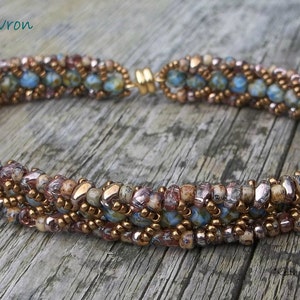 Può includere: Un braccialetto di perline con un motivo a spina di pesce. Il braccialetto presenta un mix di perline marroni, dorate e blu. La chiusura è dorata. Il testo "Mini Duo Chevron Bracelet" è visibile nell'immagine.