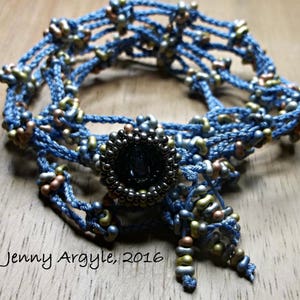 May include: A handcrafted blue beaded necklace with a central dark circular feature. The necklace showcases detailed beadwork in blue, gold, and bronze tones. The text "Jenny Argyle, 2016" is present in the image.