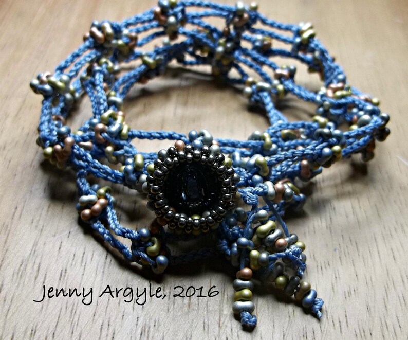 Peut inclure: Un collier de perles bleu fait &agrave; la main avec un ornement circulaire sombre au centre. Le collier pr&eacute;sente un travail de perles complexe dans des tons de bleu, d'or et de bronze. Le texte "Jenny Argyle, 2016" est visible.