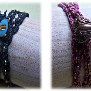May include: Two handmade bracelets. One is dark blue with gold accents and a turquoise stone. The other is purple with a decorative copper-colored clasp and beaded details. Both are displayed on a wooden surface.