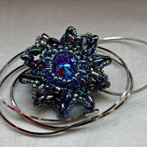 May include: A beaded pendant necklace featuring a starburst design. The pendant is primarily blue with iridescent accents and a central faceted blue gem. The necklace has a silver-colored chain.