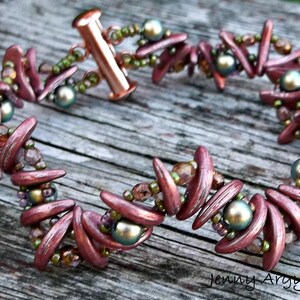 Könnte beinhalten: Ein handgefertigtes Armband mit rotbraunen, gebogenen Perlen und schillernden Akzenten. Das Armband enthält kleine grüne und schillernde Perlen sowie einen kupferfarbenen Verschluss. Das Armband wird auf einer Holzoberfläche präsentiert.