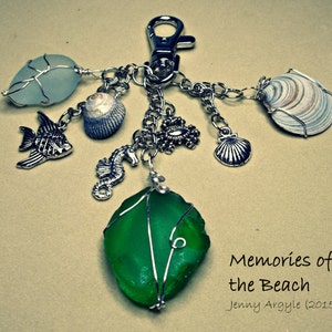 Könnte beinhalten: Ein Schlüsselanhänger mit Strandmotiven. Er zeigt ein großes grünes Seeglasstück, ein hellblaues Seeglasstück, eine Muschel, ein Seepferdchen, einen Fisch, eine Krabbe und eine kleine Jakobsmuschel. Der Text "Memories of the Beach" ist ebenfalls sichtbar.