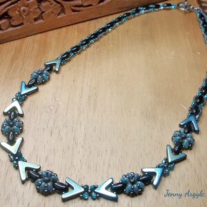 Pode incluir: Um colar de contas com detalhes em preto e verde-azulado. O colar apresenta uma s&eacute;rie de contas em forma de seta verde-azulado e pequenos grupos florais. O colar est&aacute; sobre uma superf&iacute;cie de madeira. O texto "Jenny Argyle, 2018" &eacute; vis&iacute;vel.
