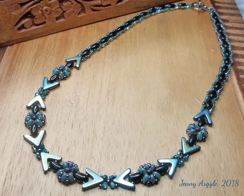 Pode incluir: Um colar de contas com detalhes em preto e verde-azulado. O colar apresenta uma s&eacute;rie de contas em forma de seta verde-azulado e pequenos grupos florais. O colar est&aacute; sobre uma superf&iacute;cie de madeira. O texto "Jenny Argyle, 2018" &eacute; vis&iacute;vel.