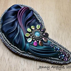 May include: A handmade brooch in a teardrop shape, with a dark blue fabric base and teal and purple detailing. It features iridescent beads, a central turquoise stone, and a silver beaded border. The text "Jenny Argyle, 2017" is visible.