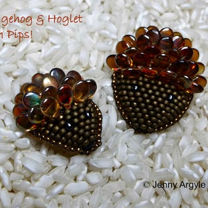 May include: Two beaded hedgehog brooches with amber and brown accents. The hedgehogs have dark eyes and are surrounded by a bed of white rice. The text "Hedgehog & Hoglet with Pips!" is at the top of the image.