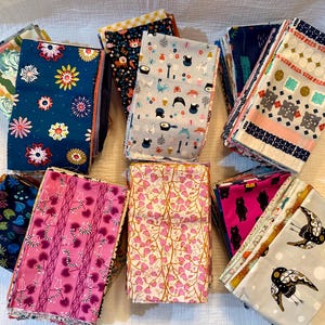 May include: A collection of folded fabric squares with various colorful floral, animal, and geometric patterns. The fabrics feature a range of colors, including blue, pink, gray, and white, and are arranged on a light-colored surface.