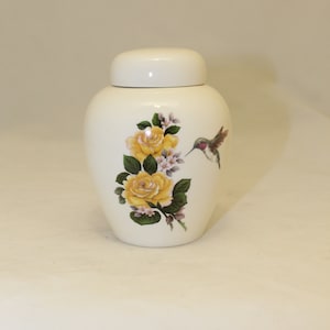 May include: White ceramic urn with a floral design featuring yellow roses and a hummingbird. The urn has a lid.