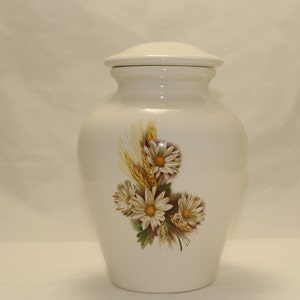 May include: White ceramic urn with a floral design featuring white daisies and wheat stalks. The urn has a lid.