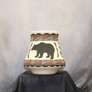 May include: A white ceramic vase with a black bear silhouette and geometric patterns in brown and black. The vase has a rounded base and a wide opening.
