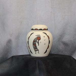 May include: A cream-colored ceramic jar with a lid, featuring a Kokopelli design in red and black. The jar is decorated with musical notes and a scalloped border. The jar is sitting on a black surface.