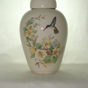 May include: A white ceramic urn with a floral design featuring yellow flowers and a hummingbird in flight. The urn has a lid and is decorated with green leaves.