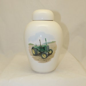 May include: A white ceramic jar with a lid, featuring a painted illustration of a green tractor with yellow wheels on a field. The jar has a rounded shape and a smooth, glossy finish.