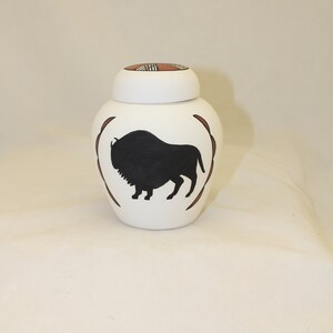 May include: White ceramic jar with a black silhouette of a buffalo on the front. The lid has a geometric design in brown and red.