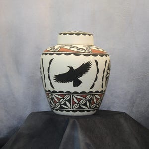 May include: A ceramic jar with a lid featuring a cream-colored base, decorated with black and terracotta geometric patterns and a central black silhouette of a bird in flight. The vessel has a rounded body and a scalloped border design.