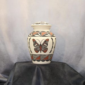 May include: Cream-colored ceramic urn with a lid, featuring a butterfly design in red, black, and white. The urn has geometric patterns around the top and bottom. The butterfly is centered on the front.
