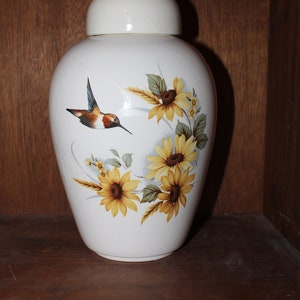 May include: White ceramic urn with a lid, featuring a painted design of a hummingbird and sunflowers. The urn has a glossy finish.