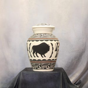 May include: Cream-colored ceramic urn with a lid, featuring a black bison silhouette and geometric patterns in black and brown. The urn is displayed on a dark surface, showcasing Native American design.