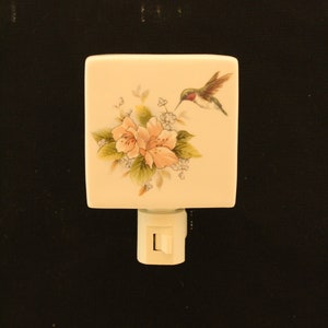 May include: A white ceramic night light with a floral design featuring pink flowers and a hummingbird. The night light has a white cord and a switch.