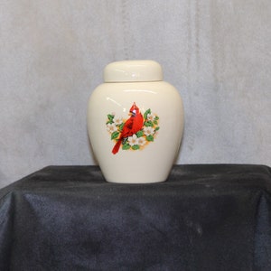May include: A white ceramic urn with a red cardinal and white flowers design. The urn has a lid.