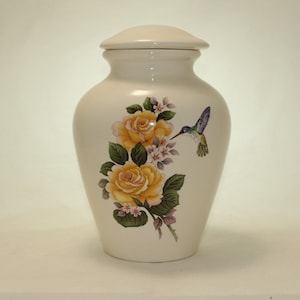 May include: Cream-colored ceramic urn with a lid, adorned with yellow roses, pink flowers, green leaves, and a hummingbird. The urn has a classic vase shape, suitable for memorial keepsakes.