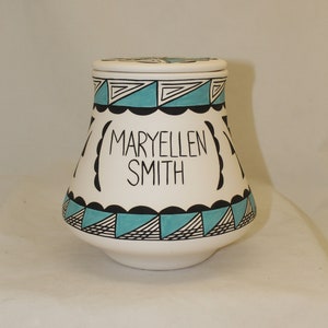 May include: White ceramic jar with a lid, featuring a turquoise and black geometric design. The jar has the name "MARYELLEN SMITH" in black lettering. The jar is a decorative item.