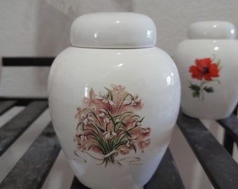 May include: White ceramic jar with a floral design. The jar has a lid and features a bouquet of white flowers with green leaves and a pink ribbon.