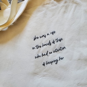 Peut inclure: Un sac fourre-tout en toile blanche avec un texte brodé en noir qui dit "She was a rose in the hands of those who had no intention of keeping her."