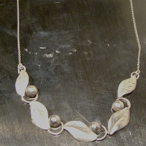 K&ouml;nnte beinhalten: Eine silberne Halskette mit einer zarten Kette und einem Anh&auml;nger aus Bl&auml;ttern und runden Perlen.